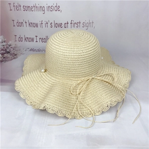 Detail of beige straw sun hat featuring wavy brim with white pearls and bow tie decoration