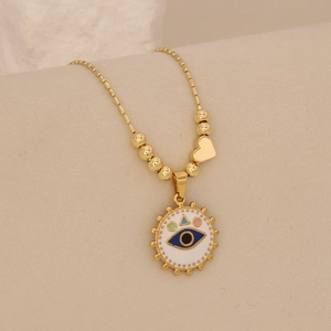 Detail of gold bead chain with white enamel evil eye pendant and heart charm on beige texture