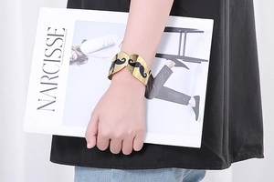 Polished gold cuff bracelet with black mustache patterns worn by a hand on a magazine
