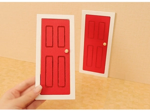 Close-up of a miniature red dollhouse door with white frame held by a hand for scale