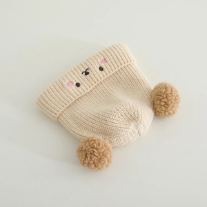 Detail of soft cream knit baby hat with furry double pom-poms and cartoon bear embroidery