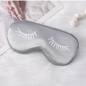 Detailed view of silver silk sleep mask with eyelash print on a soft white texture