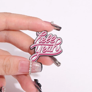 Pink and white enamel pin with 'hell YEAH' lettering held in hand for fashion accessory