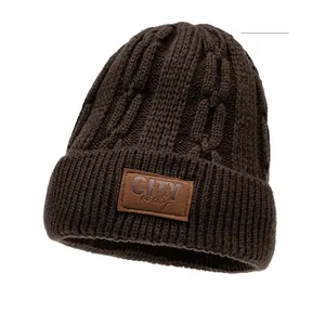Dettaglio del berretto in maglia caffè con motivo a trecce e patch logo in pelle