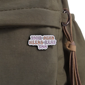 Detailed shot of an enamel pin featuring YOUR MIND NEEDS REST TOO on an olive canvas bag