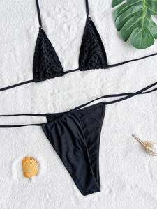 Flat lay of black triangle bikini top and mesh texture thong on white sand with palm leaf