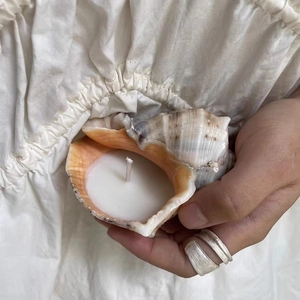 Natural conch shell scented candle in white held by a hand on beige fabric background