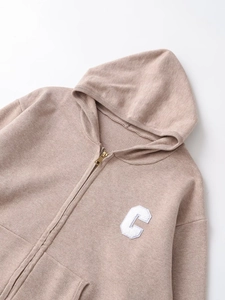 Detail of oatmeal knit hoodie featuring a towel-embroidered letter C and metal zipper