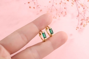 Close-up of mini gold huggie earrings with rectangular emerald green stones
