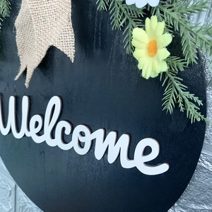 Round black wooden welcome sign with burlap bow, artificial greenery and yellow flowers