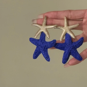 Close-up of blue and gold raffia starfish earrings held by hand for vacation style.