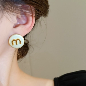 Detail of round cream enamel stud earring with gold M letter worn on model ear