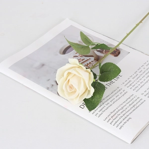Rosa artificial de terciopelo crema con hojas verdes sobre una mesa blanca