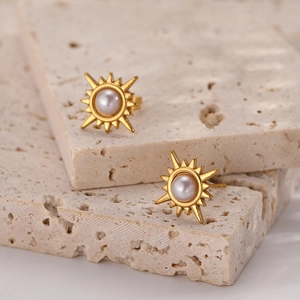 Golden sunburst stud earrings with white pearl center on a textured beige stone background