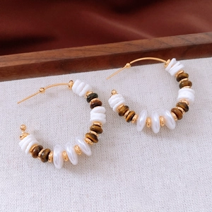 Détail de créoles dorées en C avec perles blanches et œil de tigre sur tissu blanc