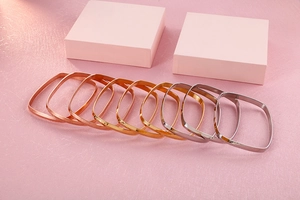 Close-up of polished square bangles in gold, rose gold, and silver finishes on pink background