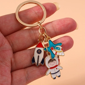 Golden keychain featuring enamel rocket, star, and astronaut charms held by a hand