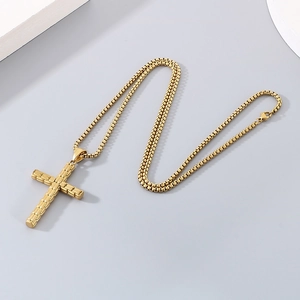 Détail d'un collier chaîne box dorée avec un pendentif croix texturé sur fond gris