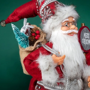 Traditional Santa Claus doll holding Merry Christmas sign and gift sack on green background
