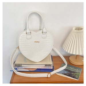 Détail du sac à main cœur blanc à motif crocodile avec logo doré posé sur des livres