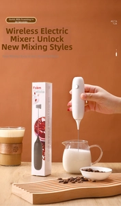 Handheld electric milk frother with metal whisk whisking milk in a glass near a coffee cup