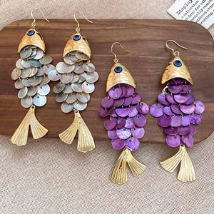 Golden fish drop earrings with shell scales and blue evil eye detail on wooden tray