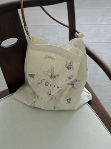 Off-white canvas shoulder bag with butterfly print and brown straps on a wooden chair