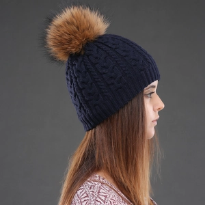 Detail of dark blue cable knit wool hat with a large brown fur pom-pom on model