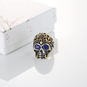 Men's skull ring in silver and gold tones with blue crystal eyes on reflective surface