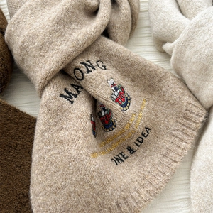 Detail eines haferfarbenen Strickschals mit Bärenstickerei und Maong-Schriftzug
