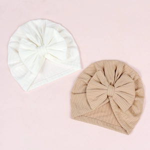 Detail of ribbed knit baby turbans with large bows in white and beige on pink background