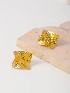 Close-up of golden wavy square stud earrings with hammered texture on beige stone