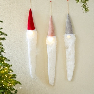 Three glowing Christmas gnome ornaments in red, pink, and grey hanging on a wall