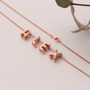 Collana con lettere ALEX in oro rosa su sfondo beige minimalista con foglie essiccate