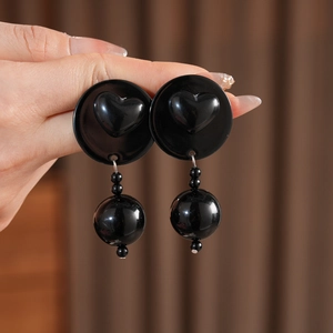 Detail view of black heart-shaped acrylic stud earrings with beaded tassels held by hand