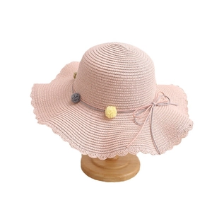 Pink kids straw sun hat with pom-poms and wavy brim on a wooden display stand