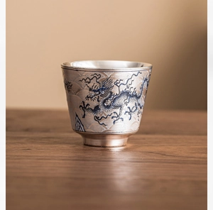 Primer plano de una taza de té plateada con motivos de dragón azul sobre mesa de madera