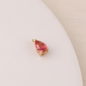 Macro detail of a pink teardrop zircon charm with gold-plated setting on a beige tray