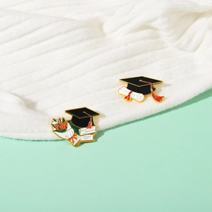 Detail view of graduation enamel pins featuring doctoral caps and books on white fabric