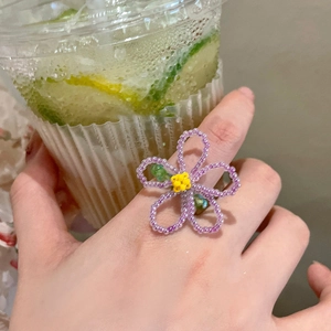 Gros plan d'une bague fleur en perles violettes avec coeur jaune et feuilles vertes au doigt