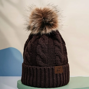 Close-up of brown knit beanie showing cable pattern, pom-pom, and leather patch