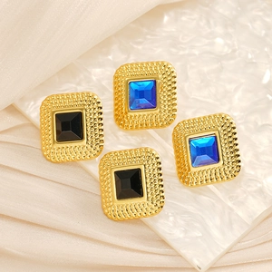 Detail view of square gold earrings with blue and black rhinestones on a white textured board