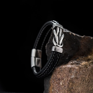 Detail of black leather bracelet with white stitching and silver knot on a dark rock