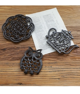 Three-piece rustic cast iron heat pads set featuring owl and teapot designs on a wooden table