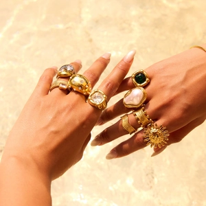 Close-up of stacked gold pearl rings with emerald cut green stones on hands by the water