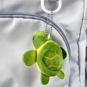 Close-up of a green plush turtle keychain with bead chain attached to a grey backpack