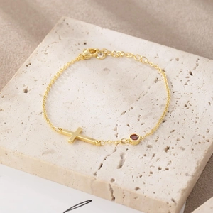 Gold minimalist cross bracelet with round red gemstone on natural stone background