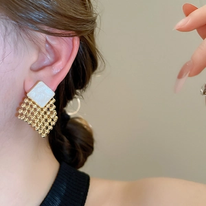 Close-up of diamond-shaped woven gold beads and white square stud earrings worn by model