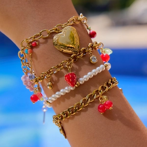Close-up of stacked gold chain bracelets with heart locket and red cherry charms