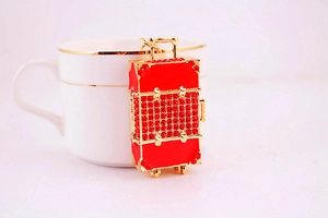 Detail of red rhinestone suitcase keychain charm with golden metal finish next to a cup